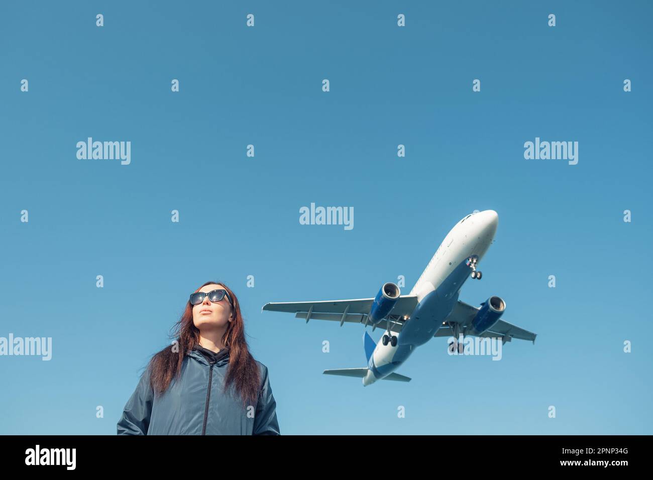 Porträt einer jungen Frau mit Sonnenbrille und einem tief fliegenden Flugzeug oder Flugzeug über ihrem Kopf gegen den blauen Himmel. Landeflugzeug über weiblichen Touristen. Trav Stockfoto