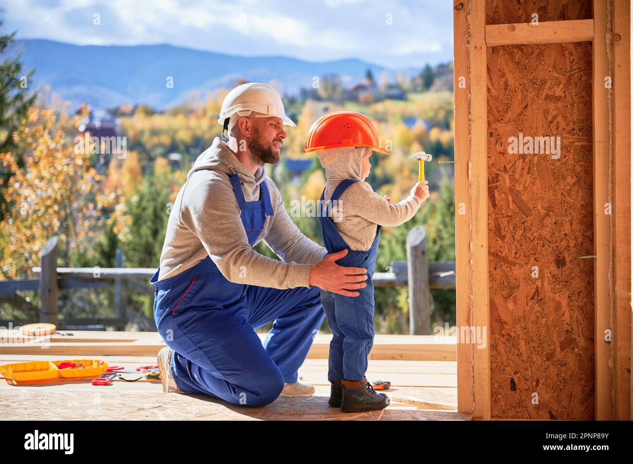 Vater mit Kleinkind Sohn Gebäude Holzrahmen Haus. Männliche Bauherren, die auf der Baustelle Nagel in die Planke schlagen, tragen an sonnigen Tagen Helm und blaue Overalls. Zimmerei- und Familienkonzept. Stockfoto