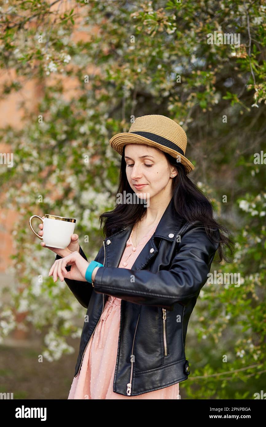 Eine Frau mit einer Tasse Kaffee neben einem blühenden Baum, schauen Sie auf die Uhr. Verspätet sein, Zeitkonzept. Lebensgleichgewicht, Geistesleben, Verbindung zum Naturpfarrer Stockfoto