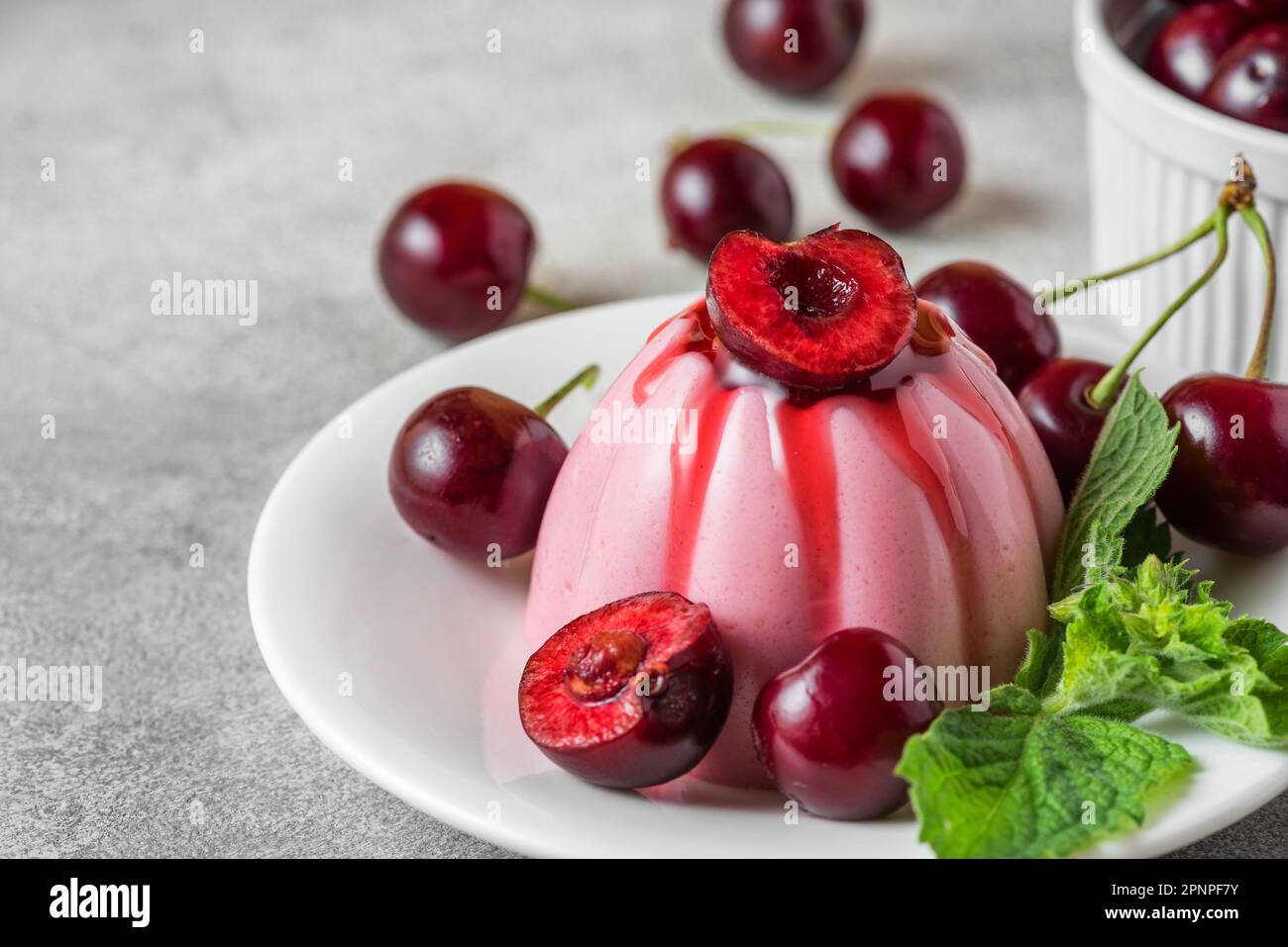 Köstliches Cherry-Panna-Cotta-Dessert mit frischen Kirschbeeren, Fruchtsauce und Minze auf einem Teller auf einem grauen Tisch. Italienisches Essen. Stockfoto
