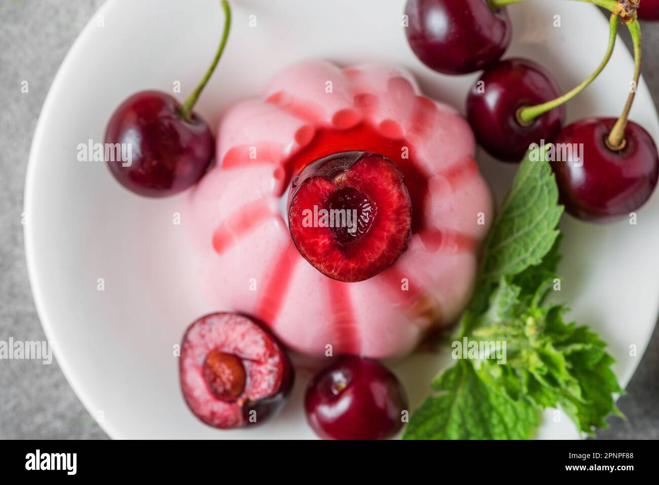 Köstliches Cherry-Panna-Cotta-Dessert mit frischen Kirschbeeren, Fruchtsauce und Minze auf einem Teller auf einem grauen Tisch. Draufsicht. Italienisches Essen. Stockfoto