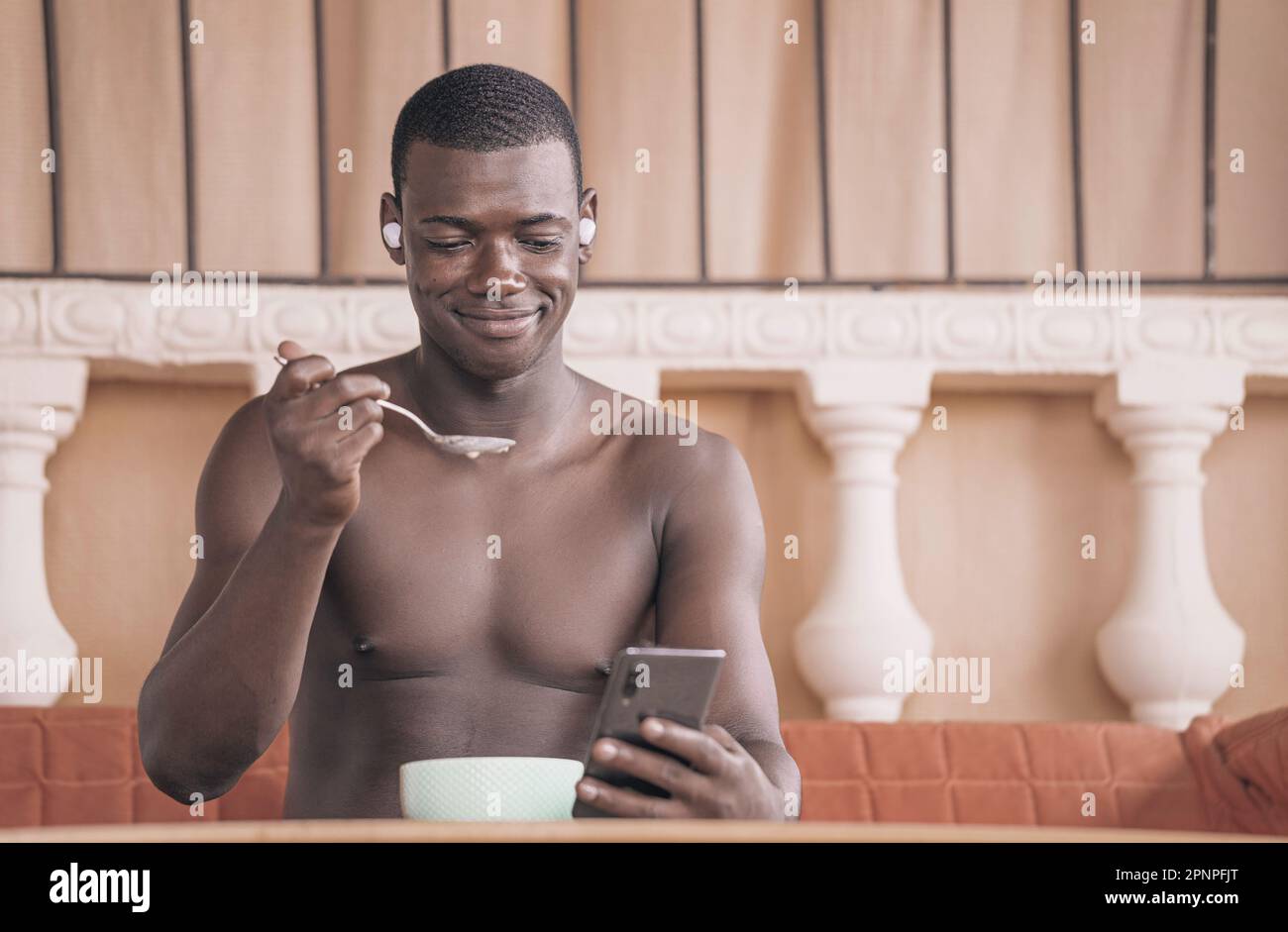 Inhalt ein afroamerikanischer Mann isst Haferbrei und liest Nachrichten auf dem Handy während des Frühstücks zu Hause. Das Bild zeigt seine Multitasking-Fähigkeiten und RE Stockfoto