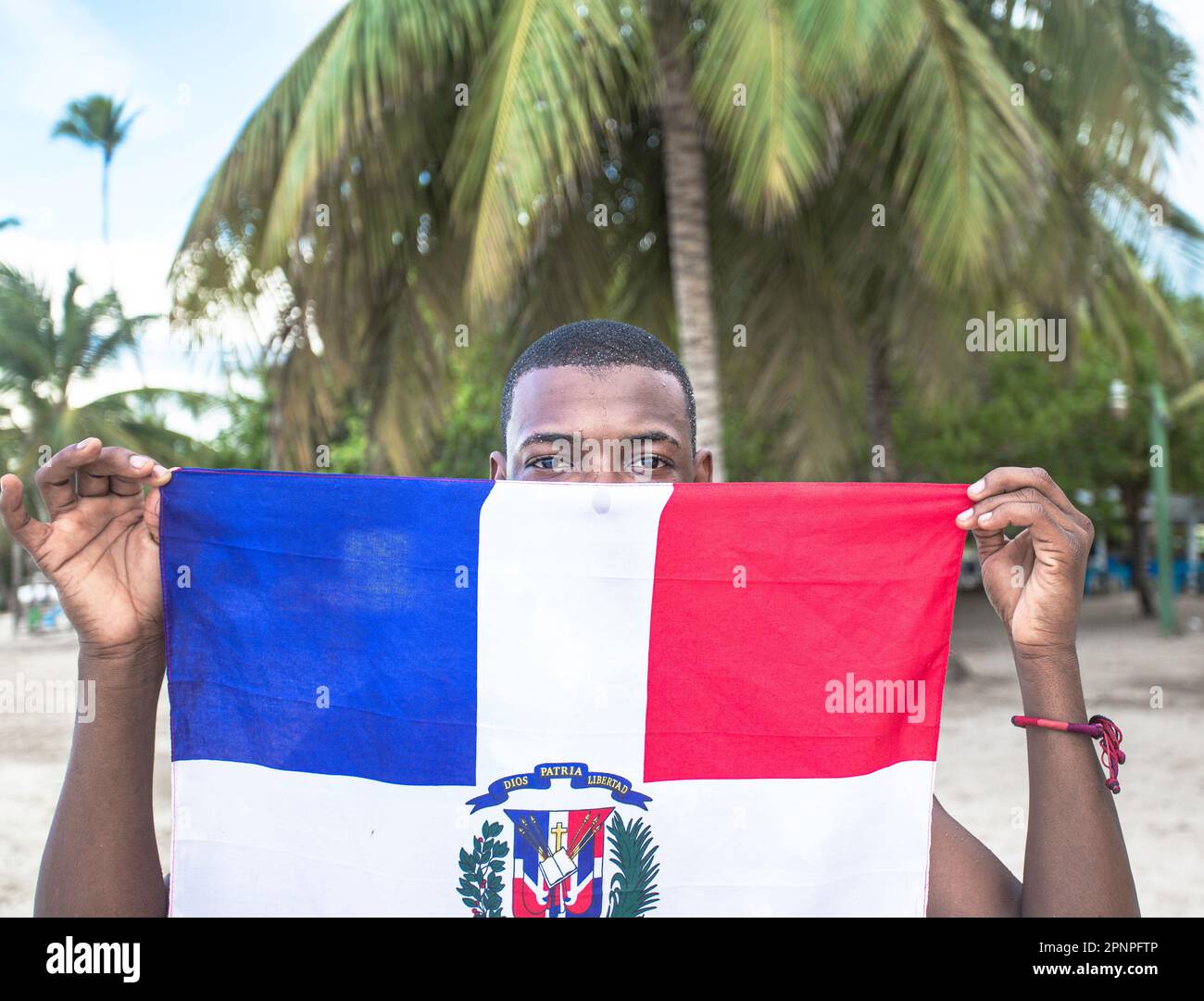 Ein stolzer kleiner schwarzer Junge zeigt die dominikanische Flagge am Strand, seine wunderschönen schwarzen Augen leuchten, während üppig grüne Palmen den Hintergrund schmücken Stockfoto