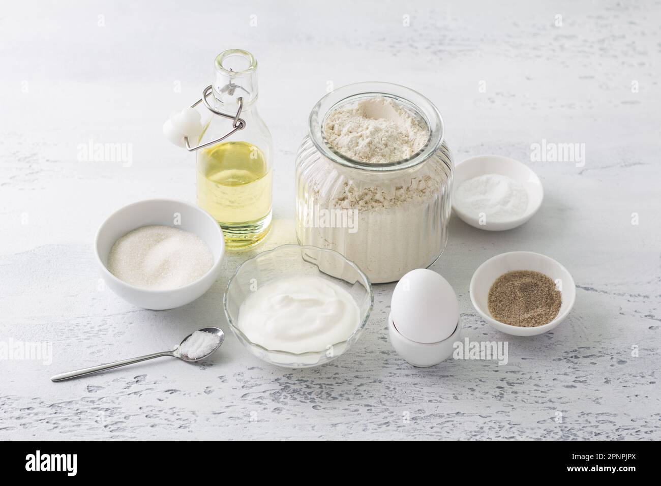 Zutaten für hausgemachtes Backen: Ei, Sauerrahm (Joghurt), Zucker, Vanillezucker, Mehl, Pflanzenöl, Backpulver, Salz auf hellblauem Hintergrund Stockfoto