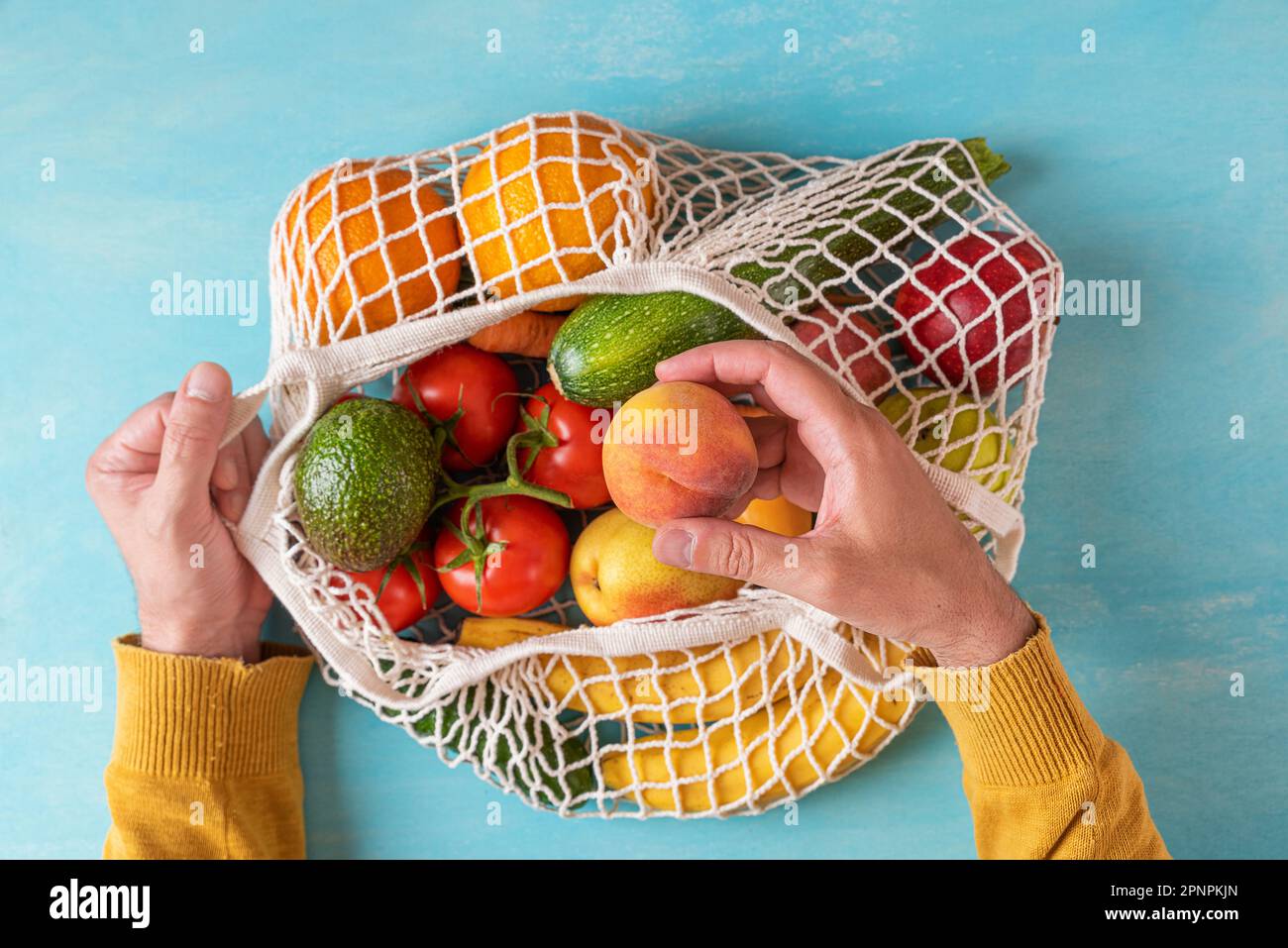 Männerhandgelenk, das Pfirsichfrucht aus einer Mesh-Einkaufstasche mit Obst und Gemüse nimmt. Umweltfreundliches Einkaufen. Nachhaltiger Lebensstil, keine Abfälle, plastikfrei Stockfoto