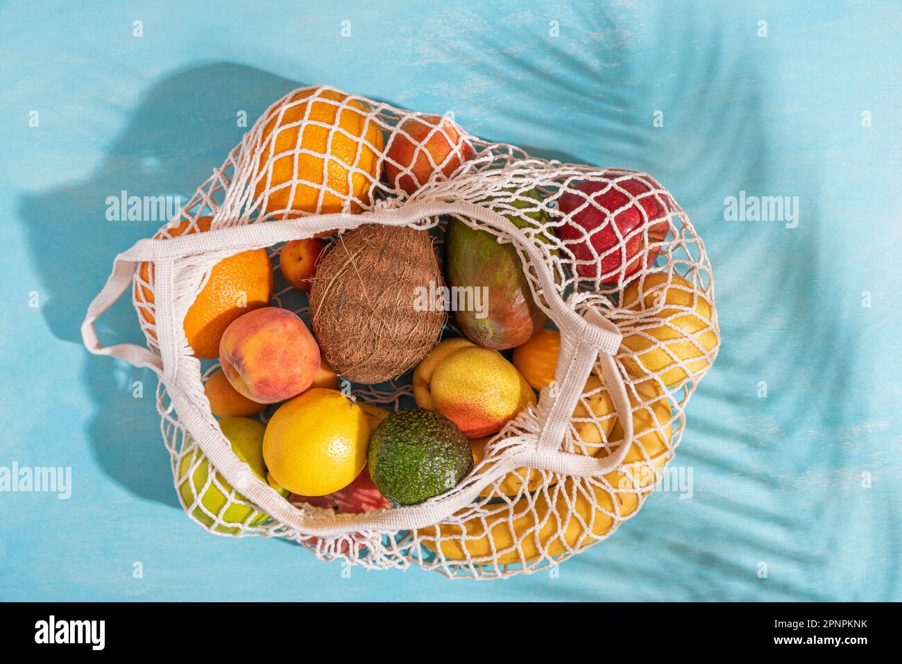 Sommerdecke mit tropischen Früchten in umweltfreundlicher Einkaufstasche. Blauer Hintergrund mit Palmenblattschatten, Sonne und Sonnenlicht. Nachhaltiger Lebensstil Stockfoto