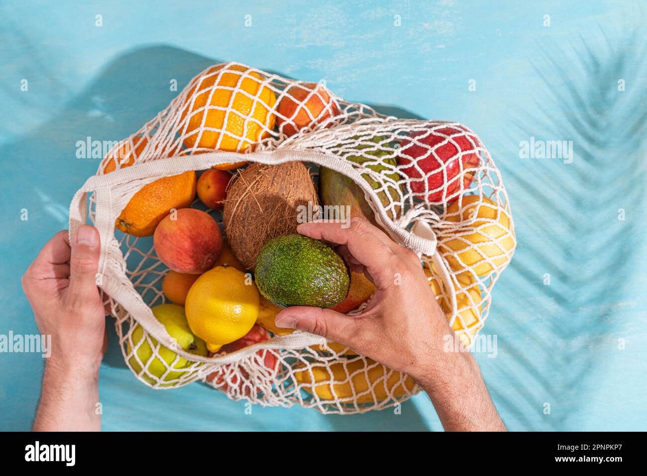 Männerhandgelenk, das Avocadofrüchte aus einer Einkaufstasche mit tropischen Früchten nimmt. Blauer Hintergrund mit Palmenblattschatten, Sonne und Sonnenlicht. Nachhaltiges Leben Stockfoto