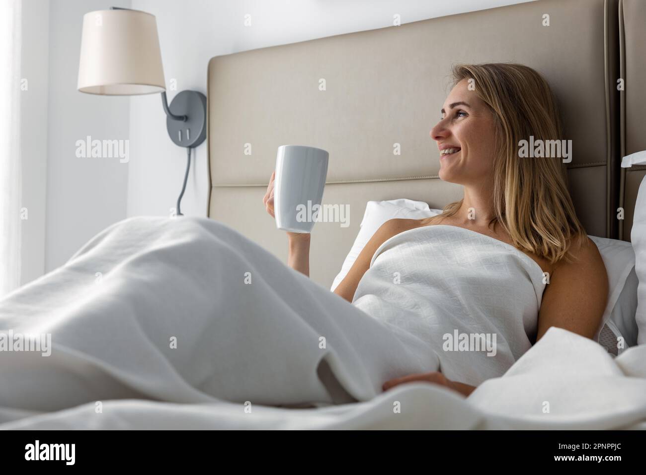 Lächelnde junge Frau, die im Bett liegt und den Morgenkaffee im Schlafzimmer genießt Stockfoto
