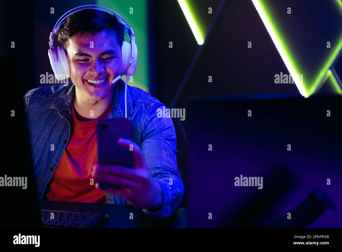 Glücklicher junger Gamer, der Live-Übertragungen von Spielen auf seinem Smartphone ansieht und streamt. Mit seinem Headset verbindet er sich und nimmt an virtuellen Spielen Teil Stockfoto