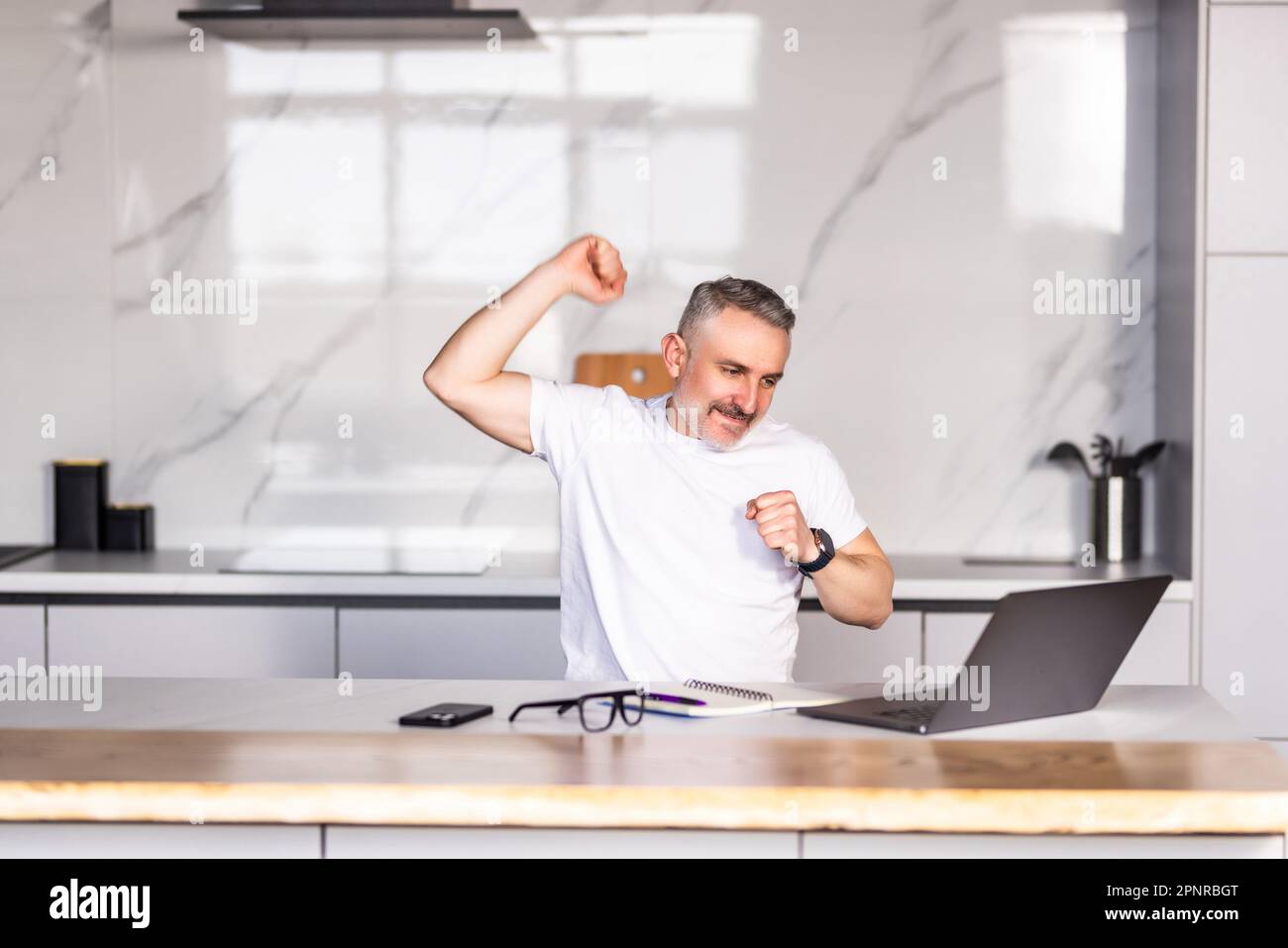 Aufgeregter Mann mit Brille sitzt am Tisch in der Küche fühlt sich euphorisch und gewinnt Online-Lotterie auf einem Laptop, überglücklich junger Mann mit Brille, schaut auf den Computerbildschirm Stockfoto