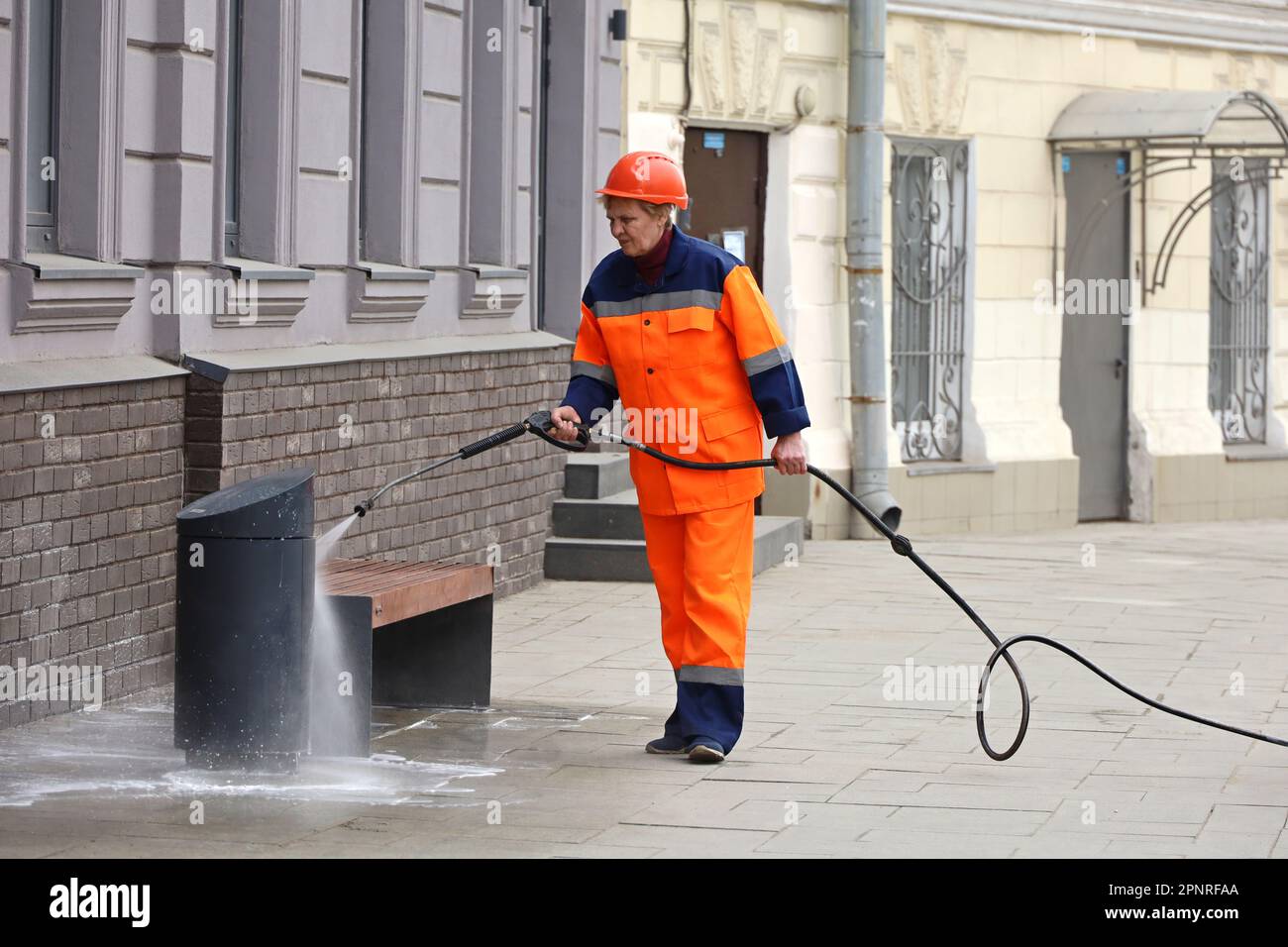 Eine Frau in Arbeiteruniform, die die Bank mit einem Schlauch bewässert. Strassenreinigung und Desinfektion in der Frühlingsstadt Stockfoto