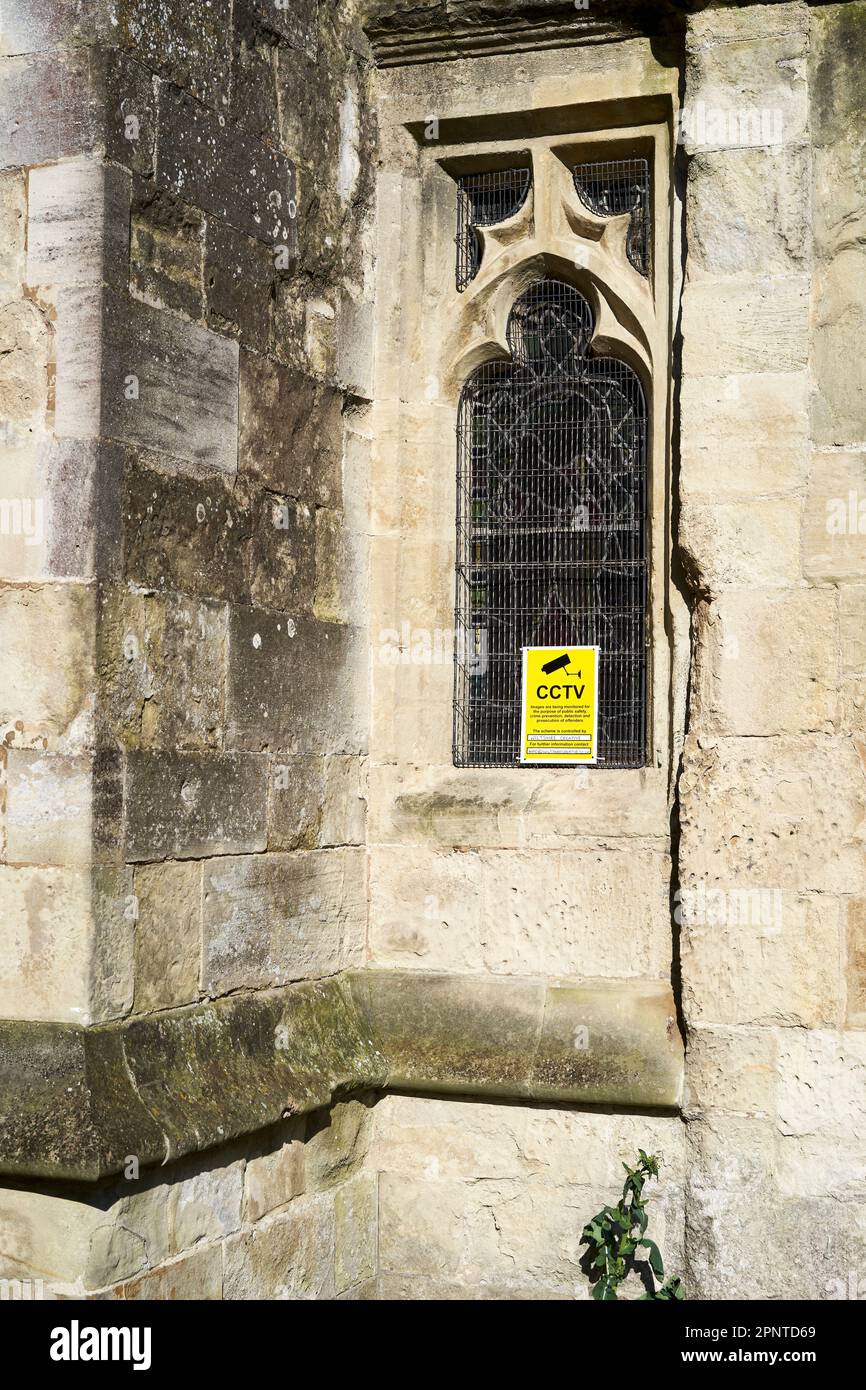 Gelber CCTV-Hinweis im bleihaltigen Fenster einer alten Steinkirche, geschützt durch einen Metallgrill Stockfoto