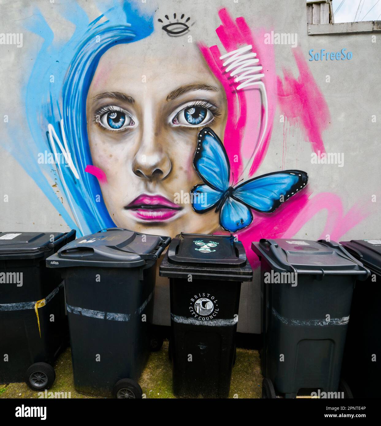 Straßenszene in Bristol UK mit Graffiti einer schönen Frau und einem blauen Schmetterling, der eine trübe Reihe von Abfalleimern erhellt Stockfoto