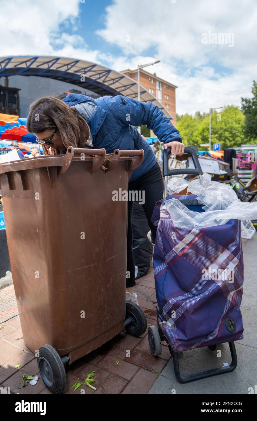 Junge Frau, die in einer Mülleimer nach Salat und Gemüse aus dem Müll mit ihrem Einkaufswagen sucht, um ihre Hühner zu recyceln und zu füttern und dabei zu helfen Stockfoto