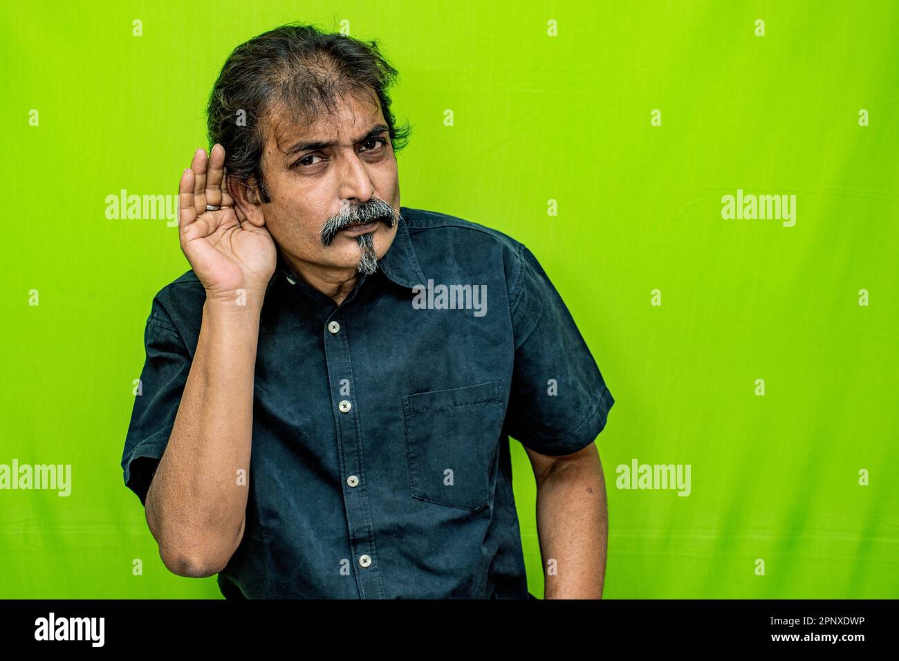 Ein Geschäftsmann in einem schwarzen Hemd steht vor einem grünen Hintergrund und schüttelt sich bewusst sein rechtes Ohr, als ob er versuchen würde, ein leises oder geheimes Geräusch zu hören Stockfoto