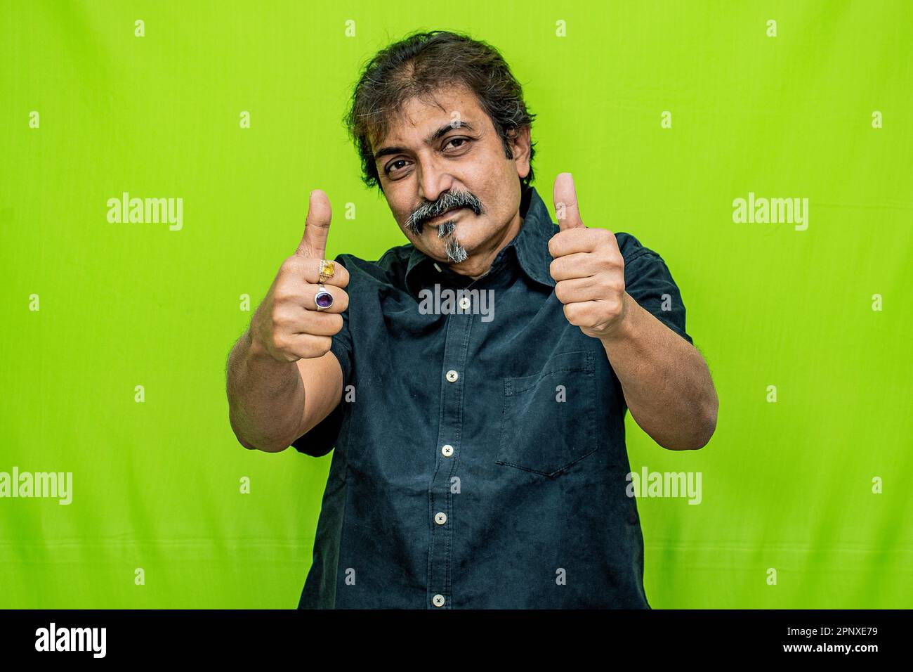 Der Geschäftsmann trägt ein schwarzes Hemd und feiert den Sieg mit erhobenen Händen und einer Daumen-hoch-Geste vor dem Hintergrund des grünen Bildschirms Stockfoto