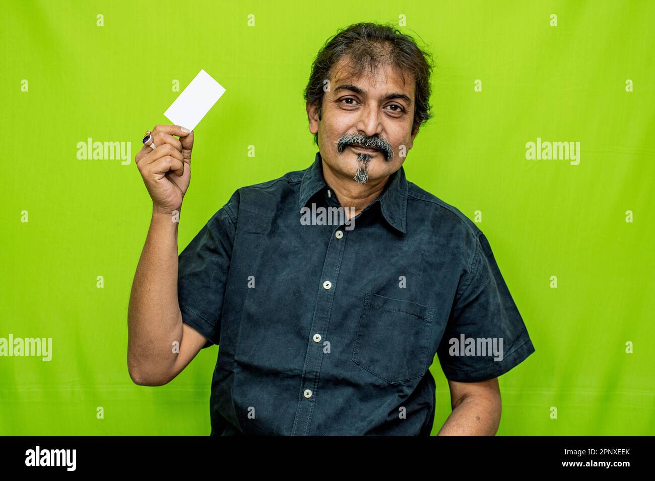 Ein lächelnder Geschäftsmann in schwarzem Hemd und einer Brille posiert mit einer leeren Kredit-/Debitkarte in der rechten Hand vor einem grünen Hintergrund Stockfoto