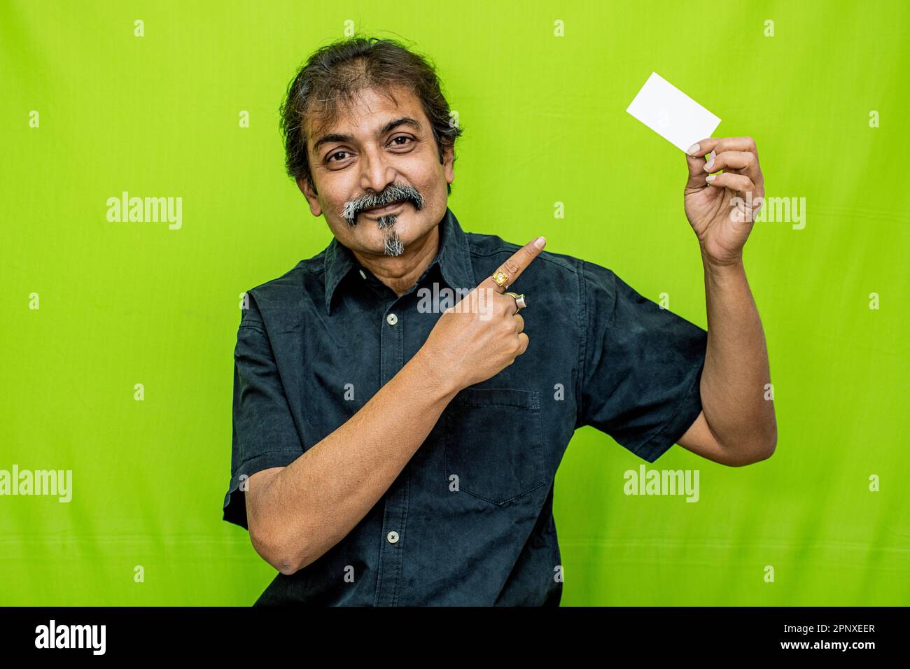 Geschäftsmann in schwarzem Hemd, lächelt mit einer leeren Karte in der linken Hand und zeigt mit der rechten auf den grünen Hintergrund Stockfoto