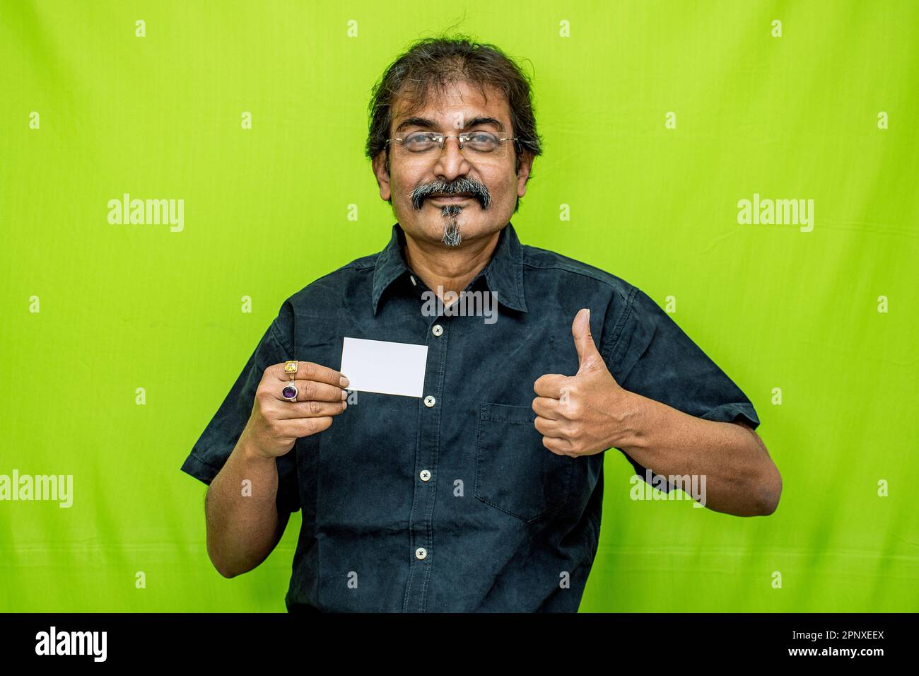 Ein Geschäftsmann in einem schwarzen Hemd und einer schwarzen Brille steht vor einem grünen Hintergrund, lächelt und hält eine Kredit-/Debitkarte in der rechten Hand Stockfoto