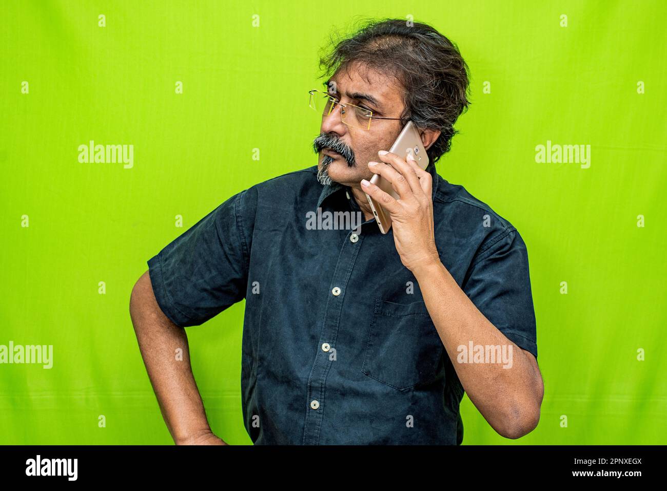 Ein gut gekleideter Indianer mit schwarzem Hemd und Brille hält sein Handy in der linken Hand und macht einen Anruf gegen einen grünen Bildschirm Stockfoto