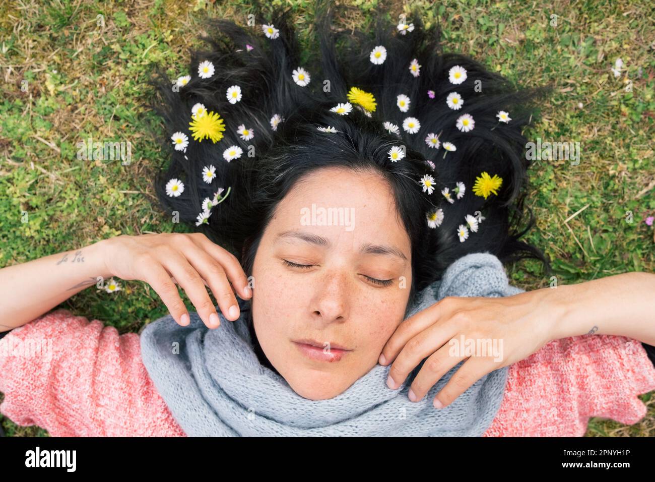 Eine Frau, die auf dem Rasen liegt, mit Blumen im Haar, Augen geschlossen und entspannt. Konzept der Frühlingsentspannung und Verbindung mit der Natur. Draufsicht. Stockfoto