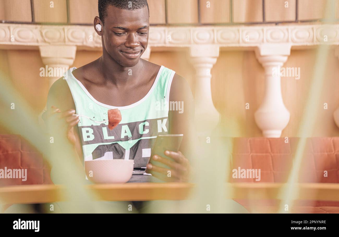 Ein lächelnder junger Schwarzer im Tank hält eine Wassermelonenscheibe auf der Gabel, während er auf der Hotelterrasse sitzt und das Telefon abfragt. Obstschale auf dem Tisch, unscharf f Stockfoto