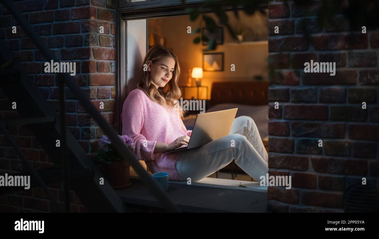 Weiße junge Frau, die nachts mit einem Notebook von Comfort of her Home auf einer Fensterbank sitzt. Weibliche Kundenberaterin, Die Dem Kundendienst Antwortet Stockfoto