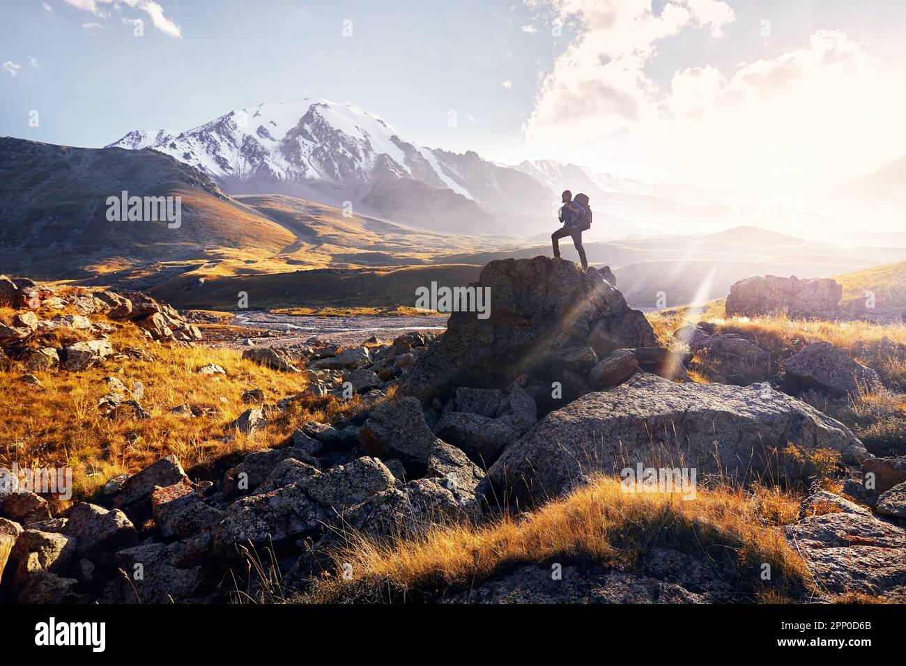 Small man Tourist mit großem Rucksack ist Wandern Klettern im Bergtal mit verschneiten Gipfeln bei Sonnenuntergang bewölkter Himmel Hintergrund in Zentralasien, Kasachst Stockfoto