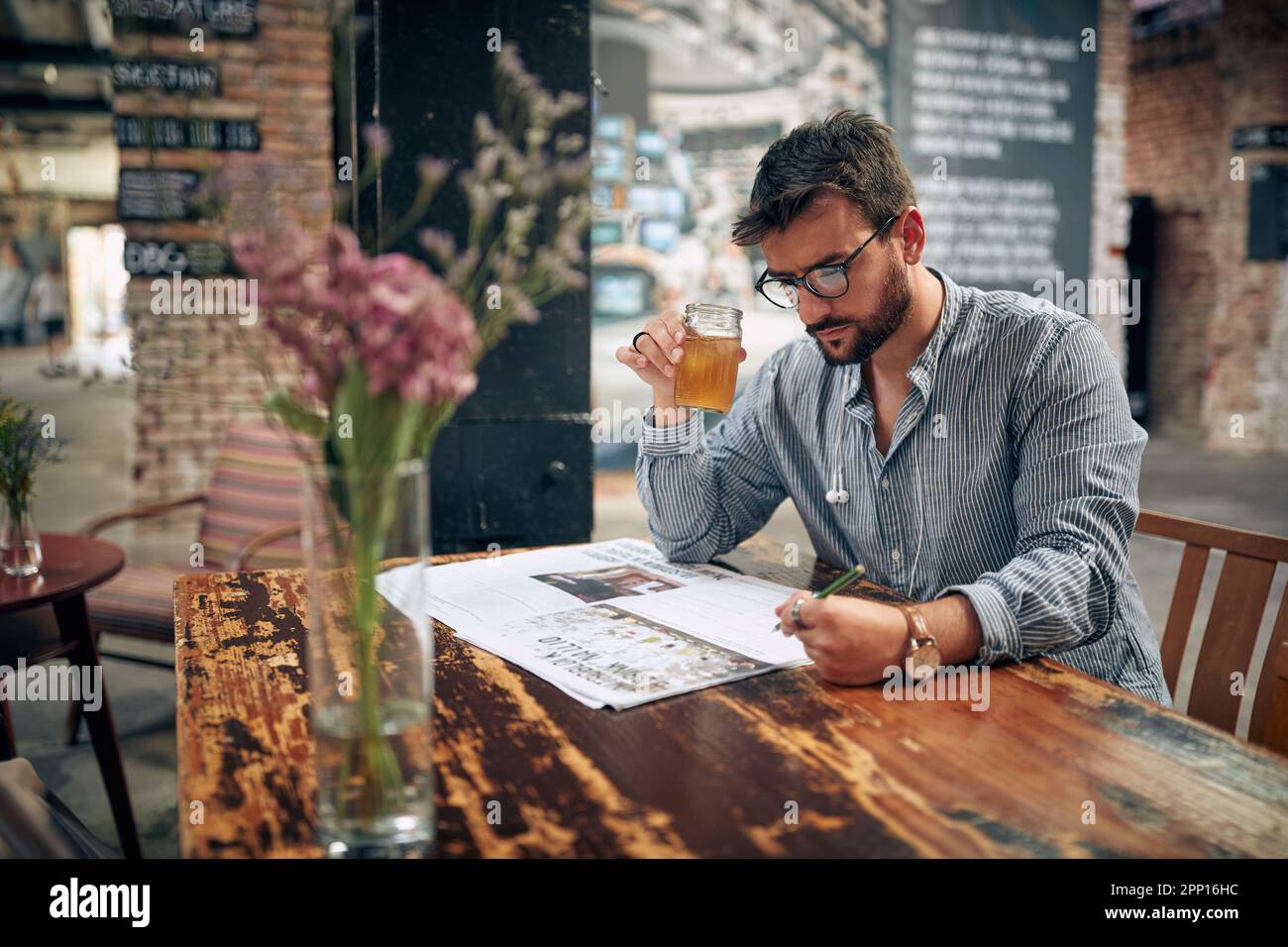 Junger Mann mit Brille und Freizeitkleidung, liest Zeitung im Café, sitzt allein. Moderne Café-Einrichtung. Stockfoto