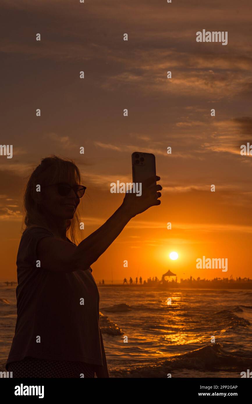 Fröhliche Frau, die Selfies mit dem Handy macht, während sie den Sonnenuntergang am Strand genießt. Stockfoto