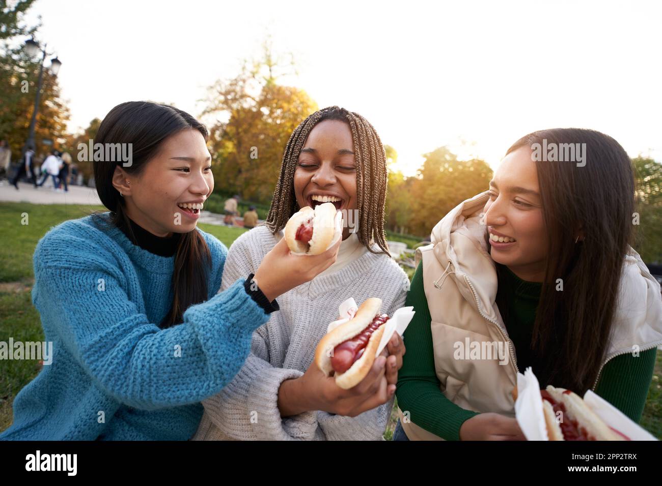 Eine Gruppe glücklicher lächelnder Mädchen, die Straßenessen essen essen und auf einer Bank in einem schönen Stadtpark sitzen. Stockfoto