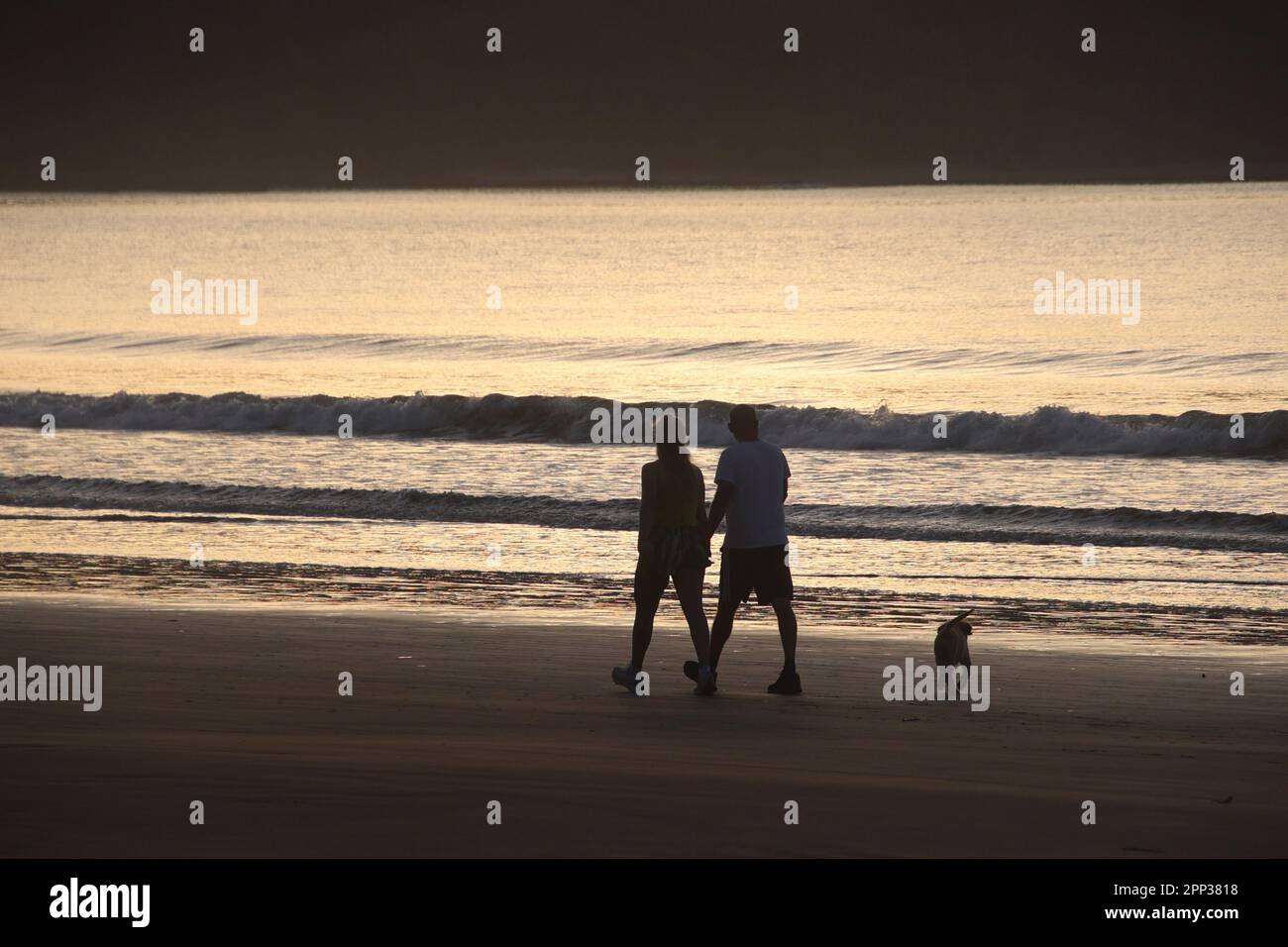 Während die Sonne über den Horizont blickt, spazieren zwei Personen mit ihrem Hundebegleiter am Strand entlang, das sanfte Wellenschlagen sorgt für einen beruhigenden Soun Stockfoto