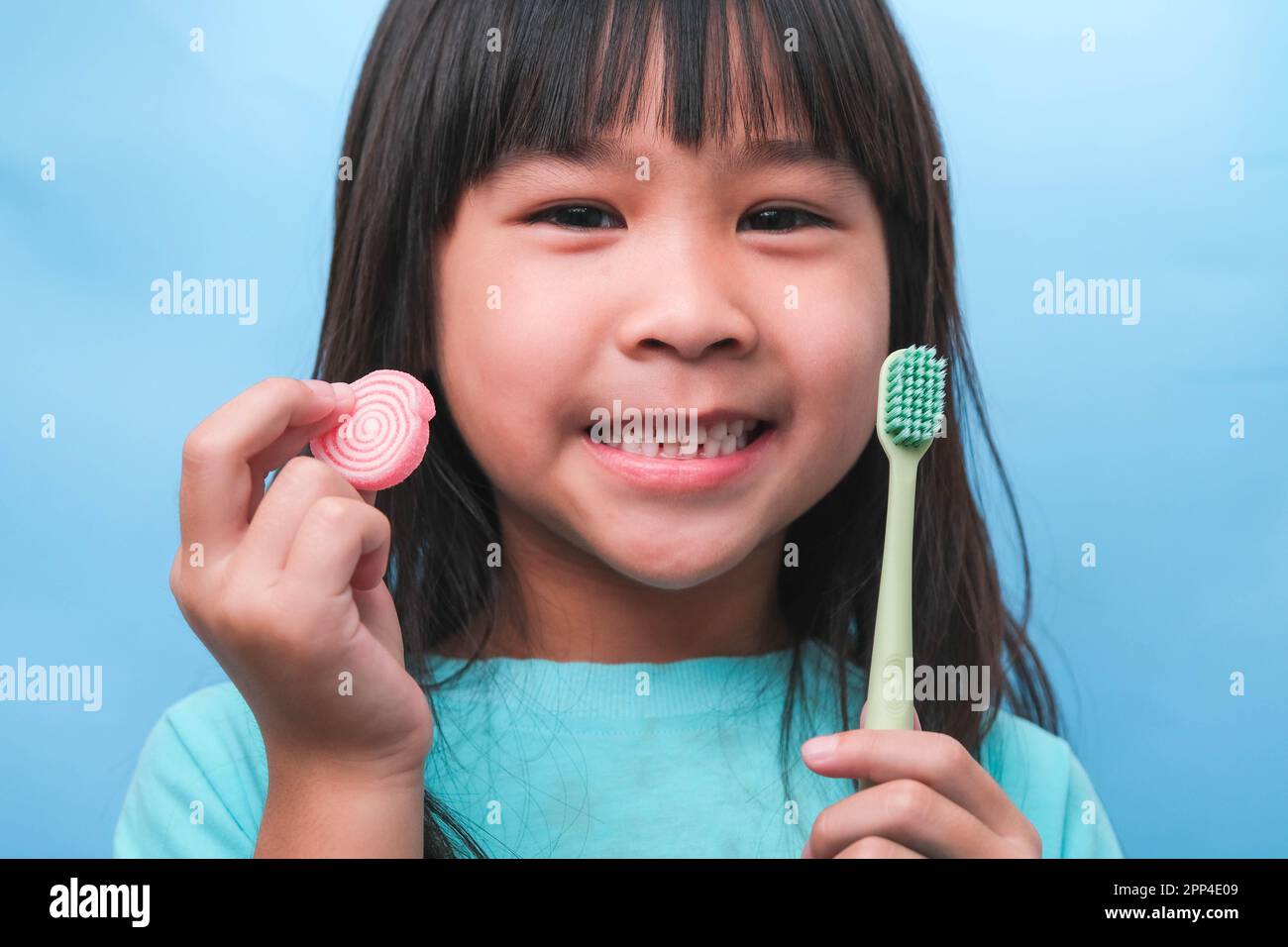 Lächelndes, süßes kleines Mädchen mit Zahnbürste und Süßigkeiten auf blauem Hintergrund. Kinder trainieren Mundhygiene und ungesunde essen. Zahnverfallsprophylaxe Stockfoto