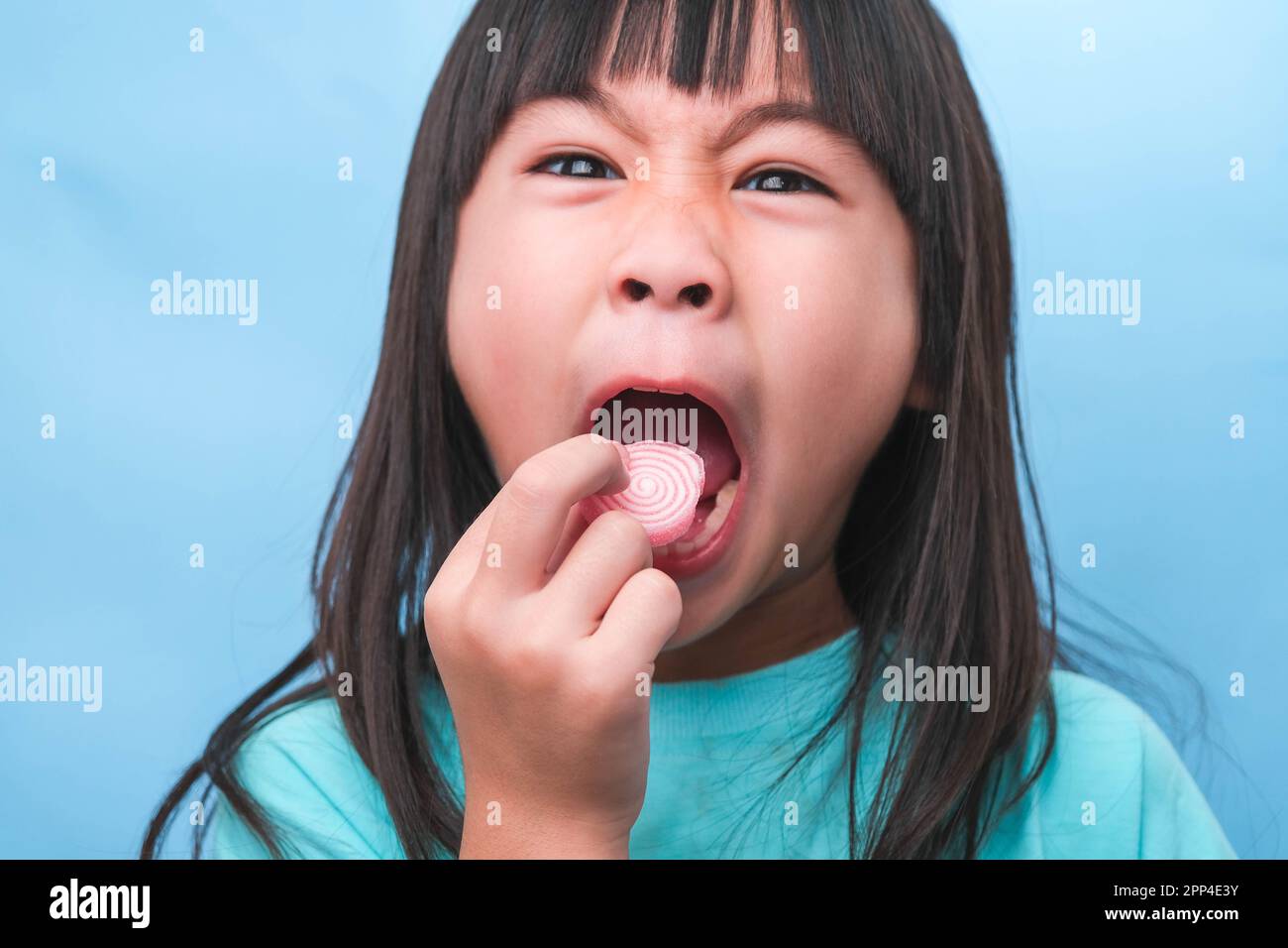 Lächelndes, süßes kleines Mädchen, das süße Gelatine mit Zucker auf blauem Hintergrund isst. Kinder essen zuckerhaltige Süßigkeiten, was zu Zahnverlust oder Zahnverlust führt Stockfoto