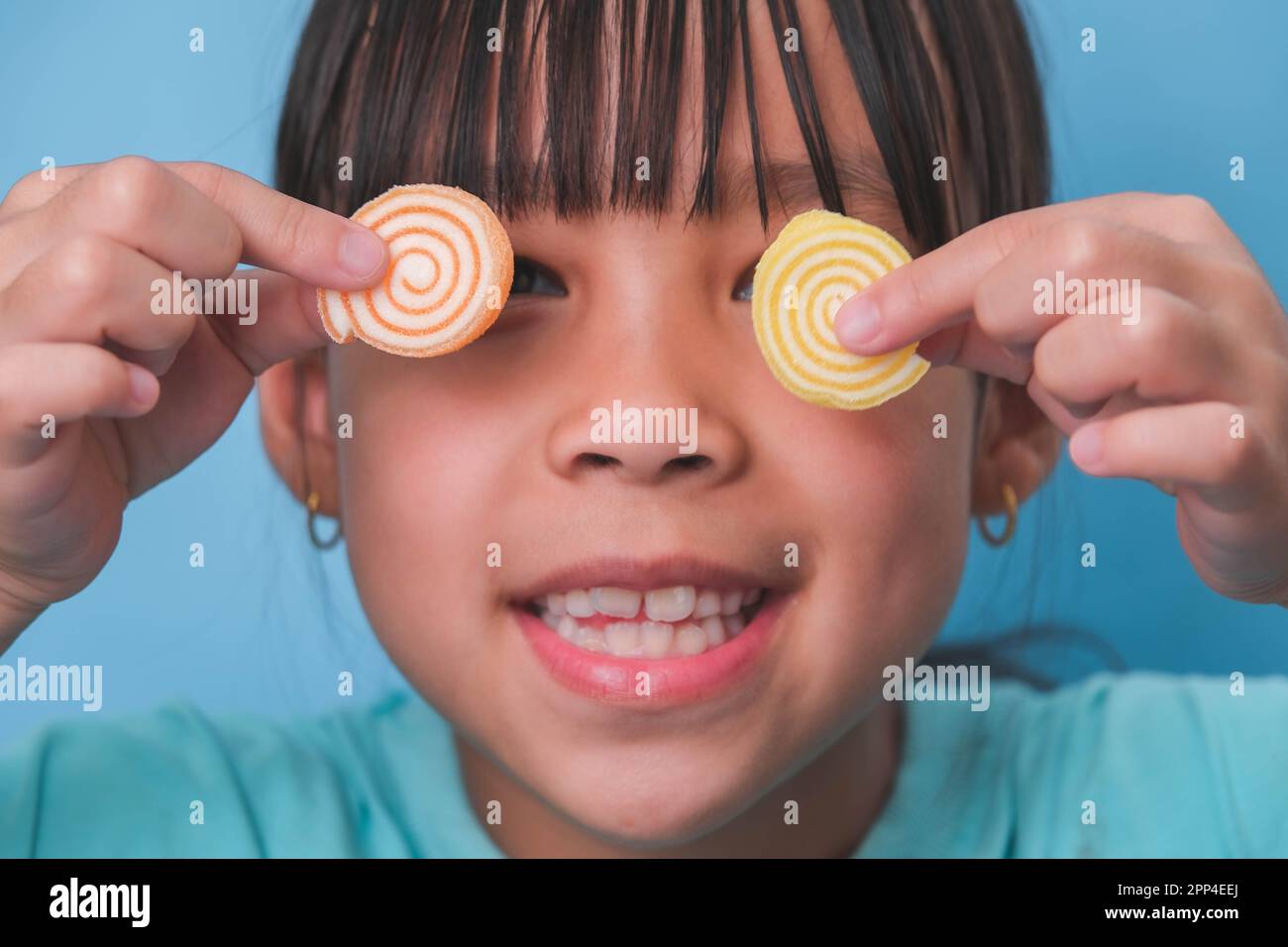Lächelndes, süßes Mädchen, das seine Augen mit zwei Gelatinesüßen auf blauem Hintergrund bedeckte. Kinder essen zuckerhaltige Süßigkeiten, die Zahnverluste verursachen oder Stockfoto
