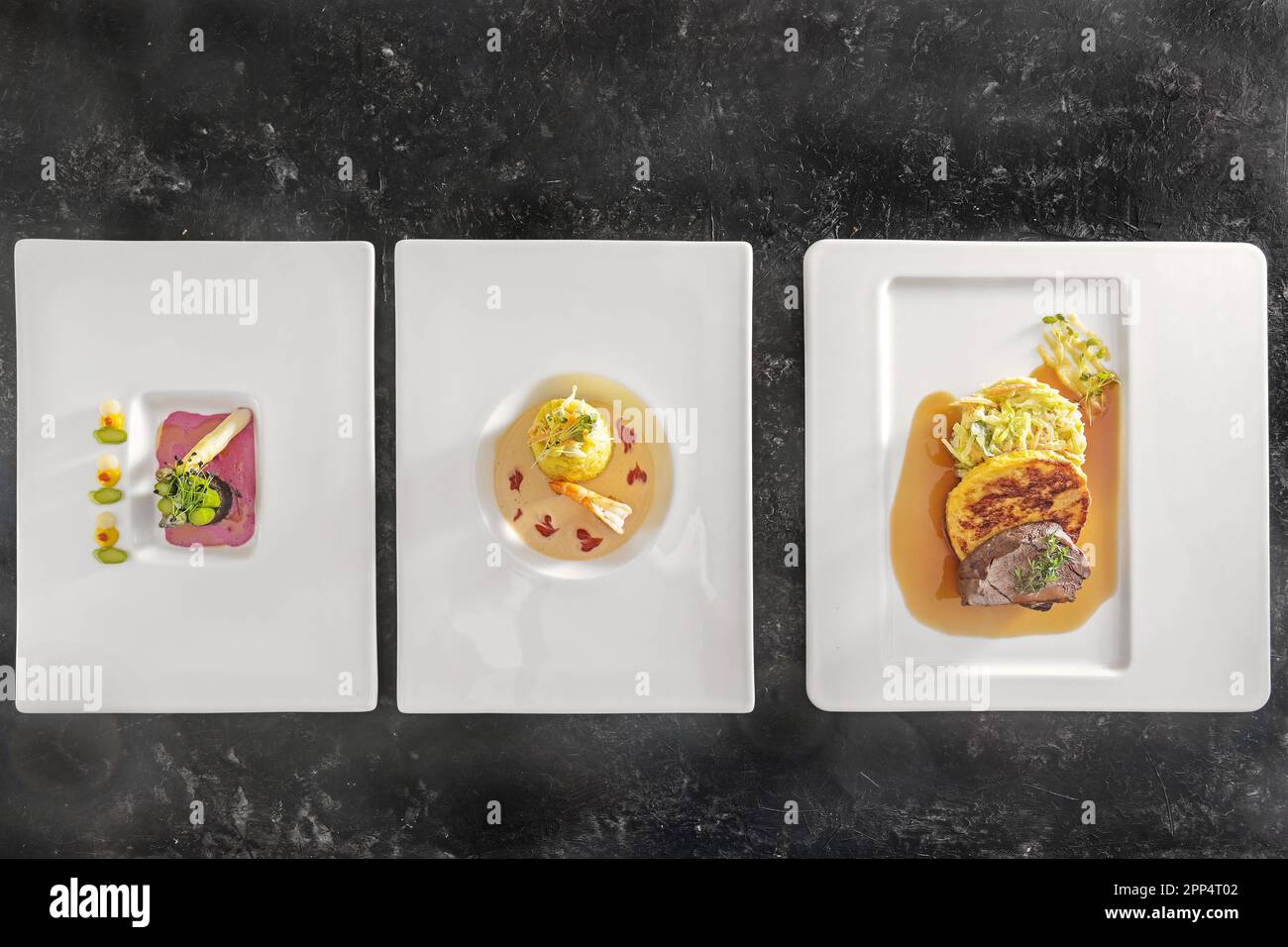 Drei Gerichte auf weißen Tellern, Spargel mit Rote-Bete-Sauce, Safran-Risotto mit Garnelen, geröstetes Hirsch mit Teigtaschen und Gemüse, dunkler Hintergrund, Stockfoto