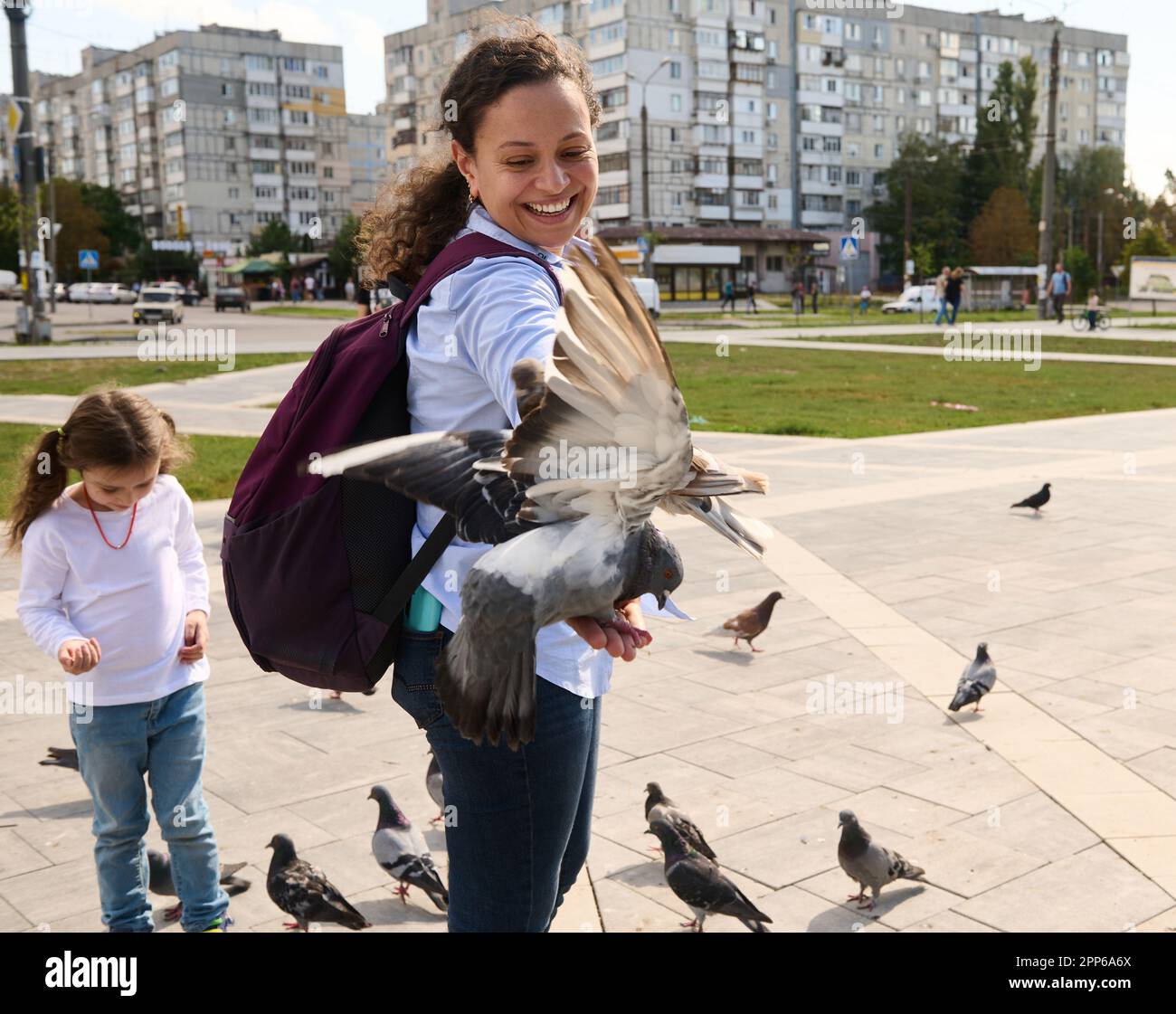 Lächelnde, multiethnische Touristinnen mit Rucksack, die positive Emotionen ausdrücken, während sie Felsentauben und wilde Tauben auf dem städtischen Platz füttern. Der Betrug Stockfoto