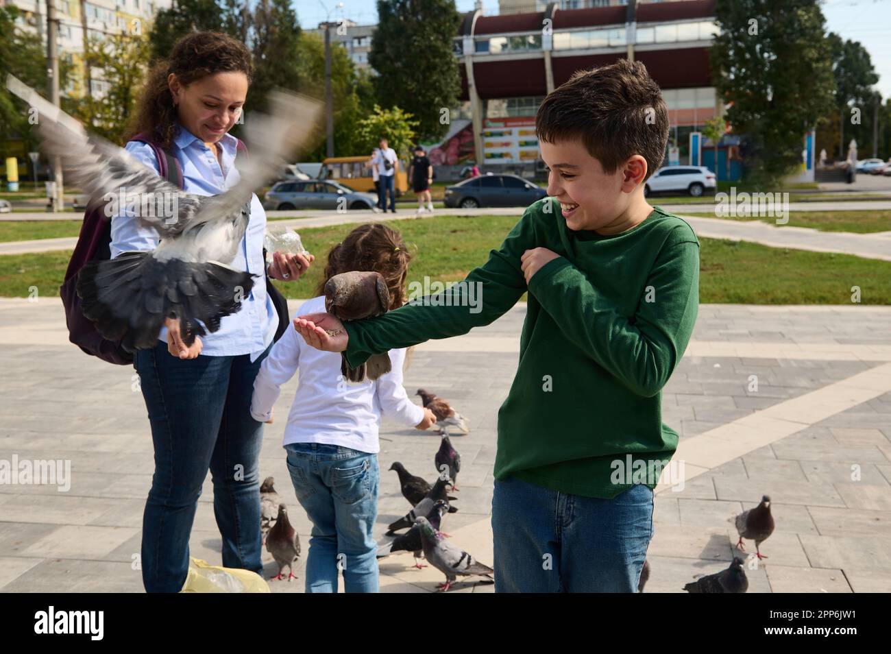 Glückliche, multiethnische Familie mit liebevoller Mutter und zwei Kindern, die Tauben auf dem Platz eines Stadtparks füttern. Menschen. Die Natur. Menschliche Beziehungen. Familienunterkünfte Stockfoto