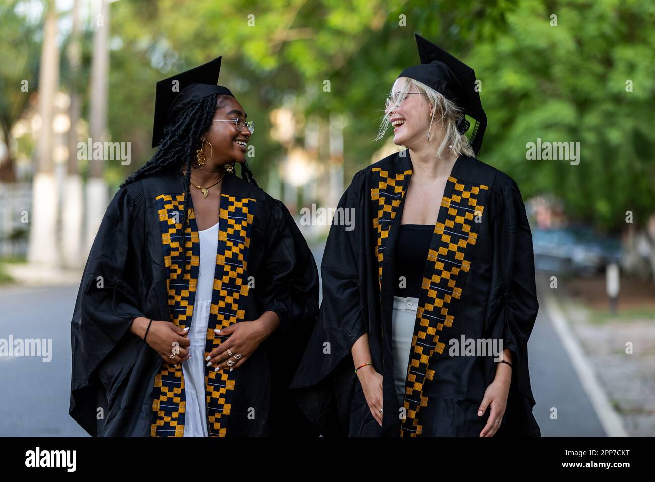 Multiethnische, schwarze und kaukasische weibliche Universitätsabsolventen tragen Kittel und Mützen und teilen auf dem Weg zur Abschlussfeier einen Witz. Stockfoto