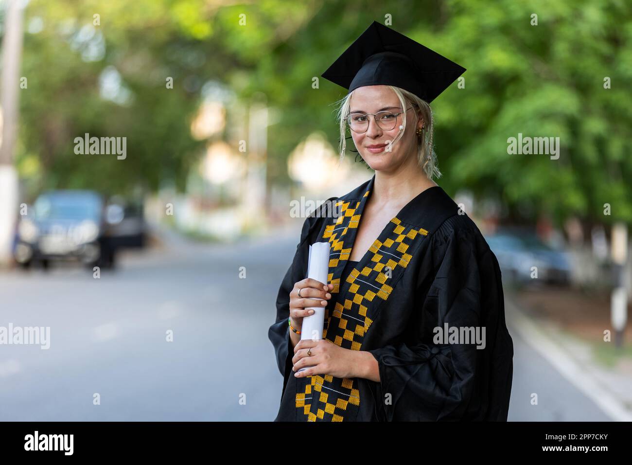 Porträt einer weiß-kaukasischen weiblichen Universitätsabsolventin mit schwarzem Kleid und Mütze, mit ihrem Abschlusszeugnis, stolz auf ihre akademische Leistung, erfolgreich Stockfoto