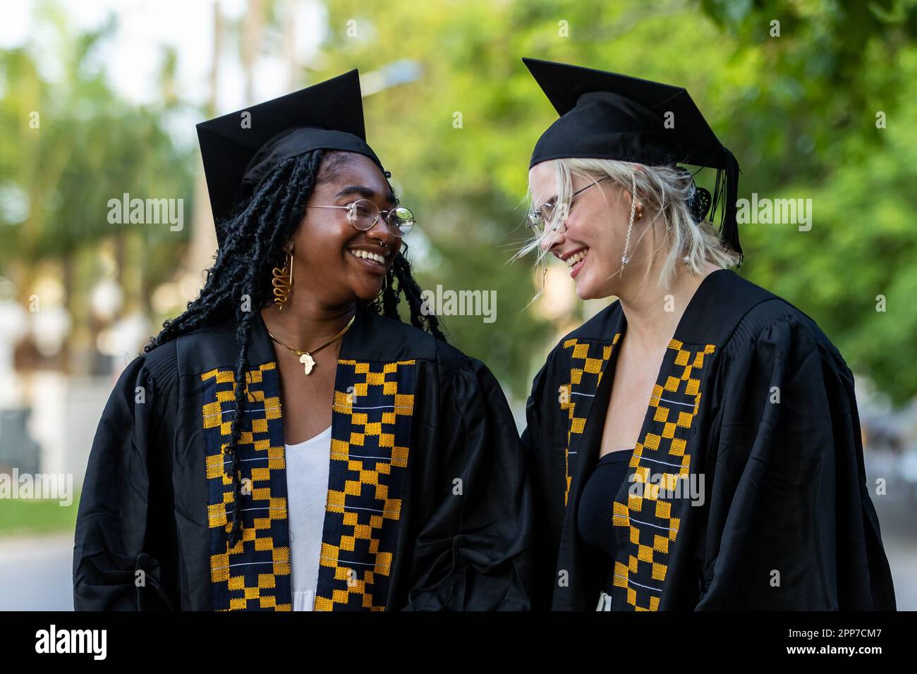 Schwarzafrikanische und kaukasische weibliche College-Absolventen, die Kappen und Kleider tragen, lachen auf dem Campus, sind stolz auf akademischen Erfolg, Diplome und das Erreichen von k Stockfoto