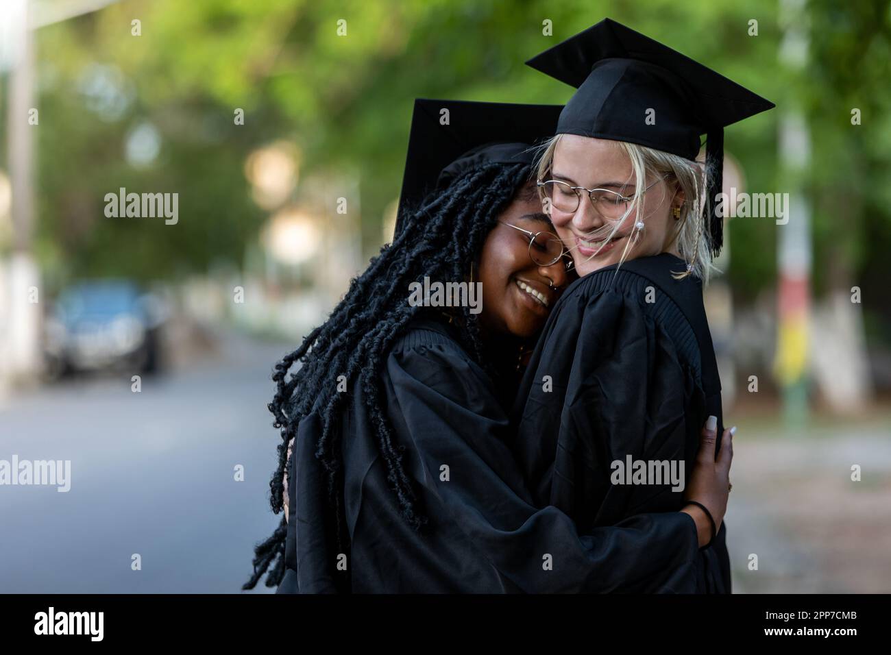 Schwarzafrikanische und kaukasische weibliche College-Absolventen, die Kappen und Kittel tragen, umarmen sich nach dem Abschluss, sind stolz auf akademischen Erfolg, Abschlüsse und gewinnen Wissen Stockfoto