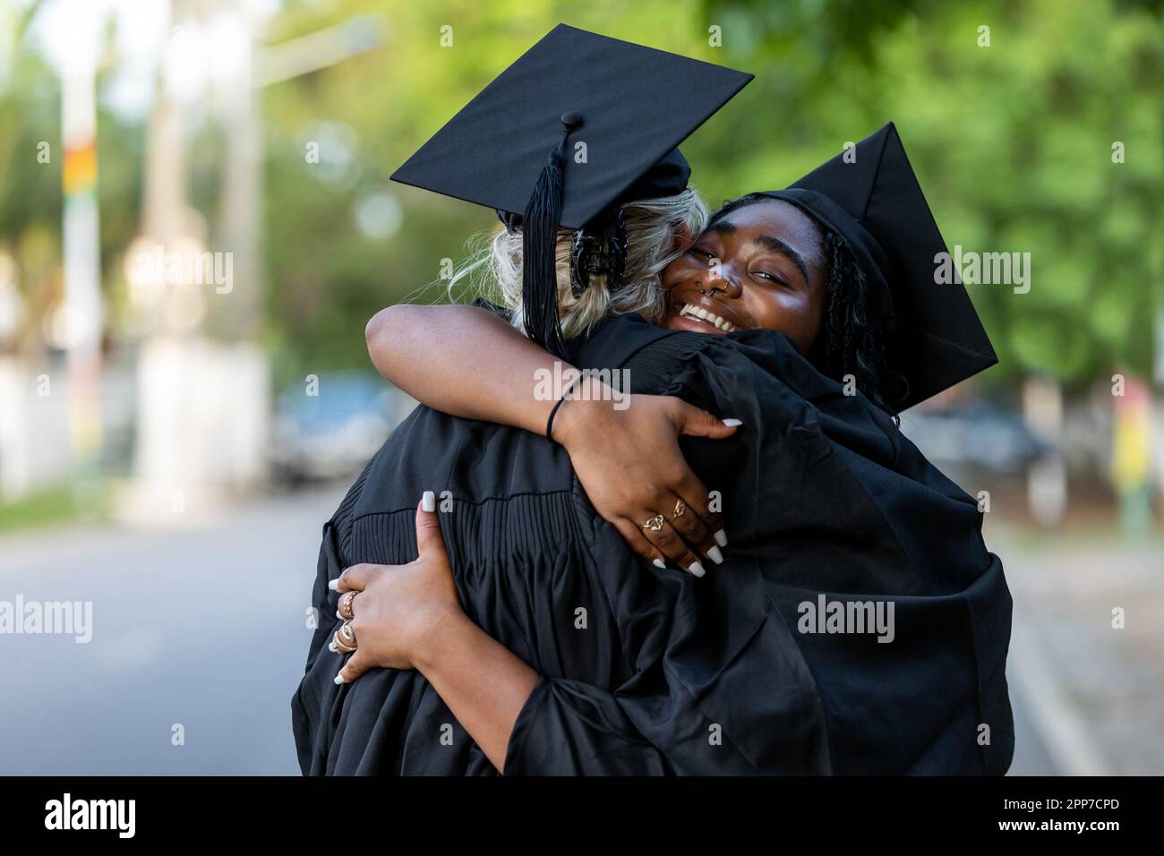 Schwarze und kaukasische weibliche College-Absolventen, die Kappen und Kittel tragen, umarmen sich nach dem Abschluss, sind stolz auf akademischen Erfolg, Abschlüsse und das Erlernen von Kenntnissen Stockfoto