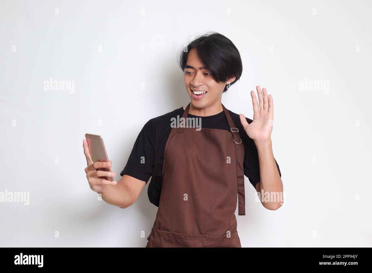 Porträt eines attraktiven asiatischen Baristas in einer braunen Schürze, der während eines Videogesprächs eine gute Geste macht. Isoliertes Bild auf weißem Hintergrund Stockfoto