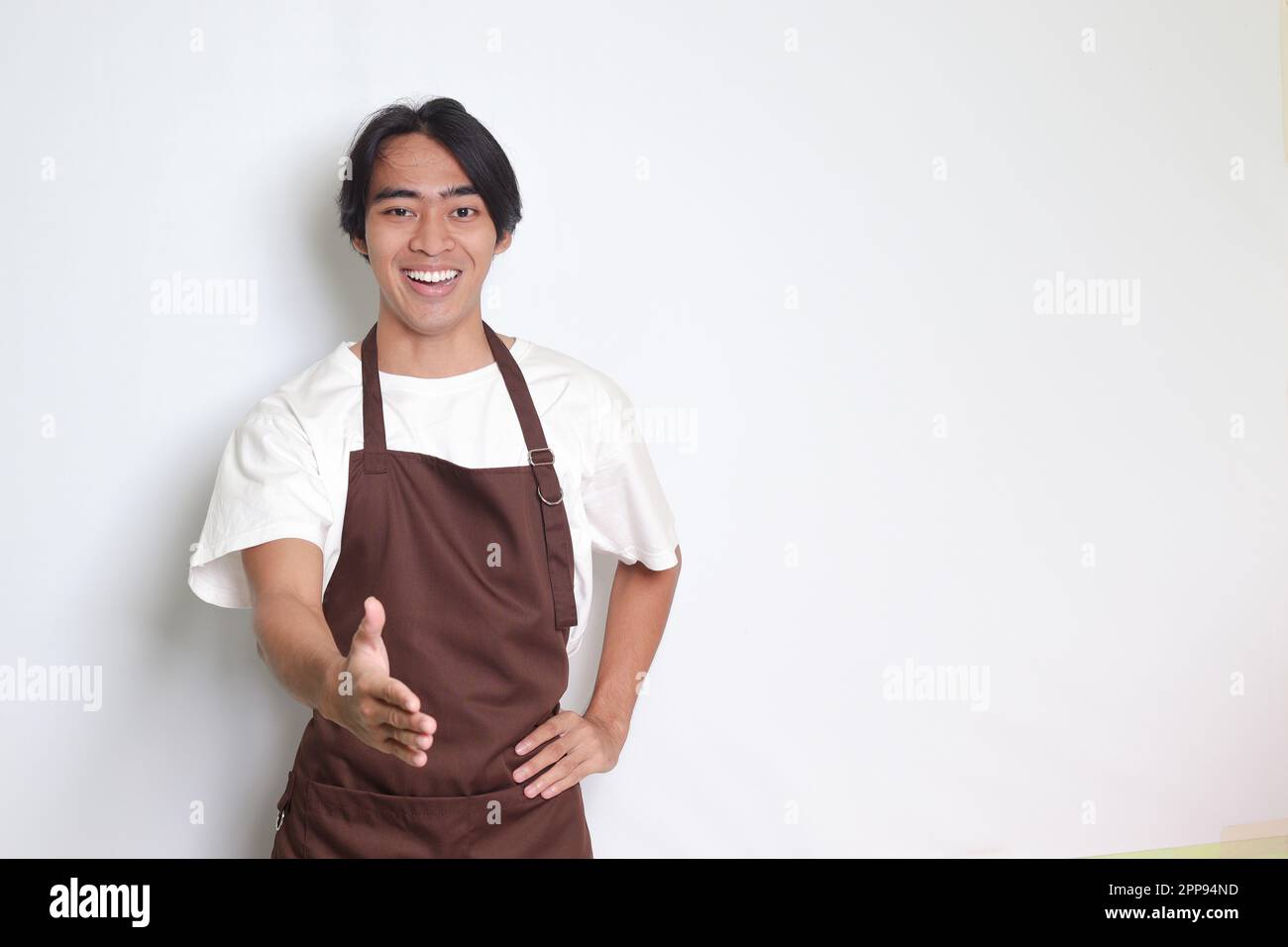 Porträt eines attraktiven asiatischen Barista in brauner Schürze, der nach einem Händedruck streckt und jemanden begrüßt. Isoliertes Bild auf weißem Hintergrund Stockfoto