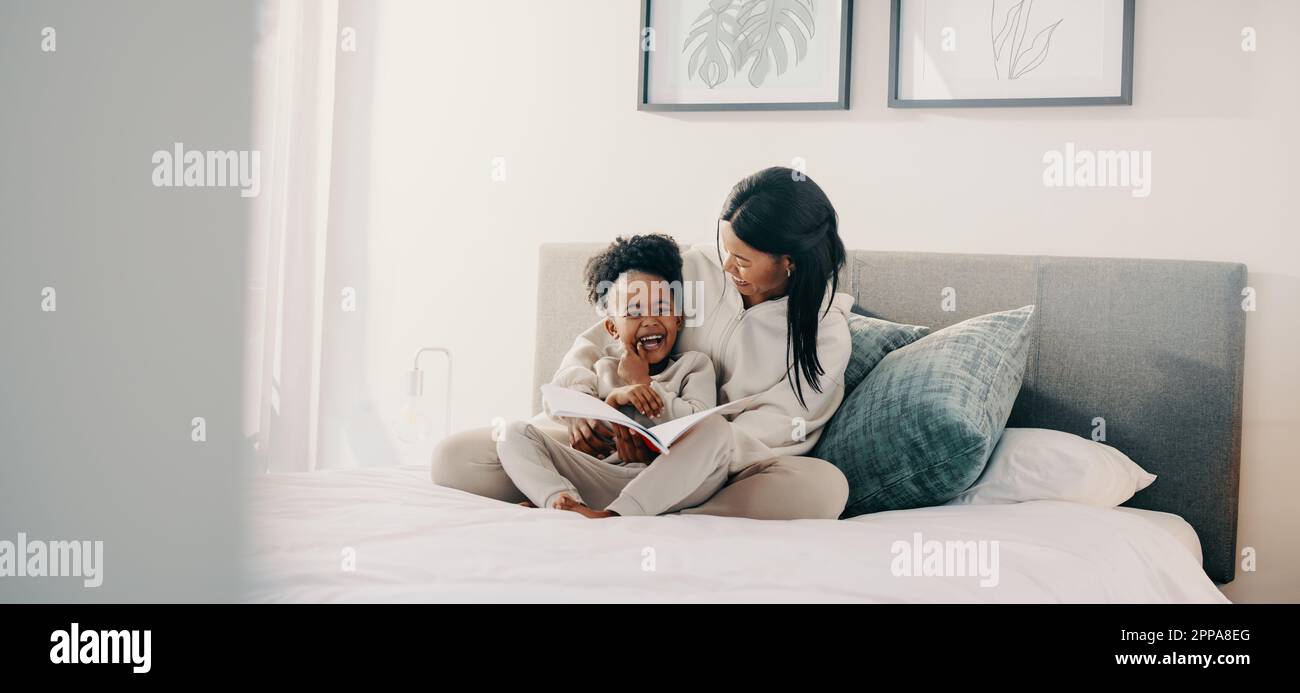 Mom und Tochter lachten zusammen, während sie ein Geschichtenbuch auf einem Bett lasen. Ein junges Mädchen im Grundalter wird von einem Märchen unterhalten, das sie liest Stockfoto