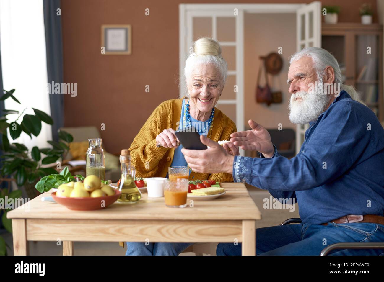 Ein älterer Mann zeigte auf den Bildschirm des Smartphones und sprach mit seiner Frau während des Frühstücks zu Hause am Tisch Stockfoto