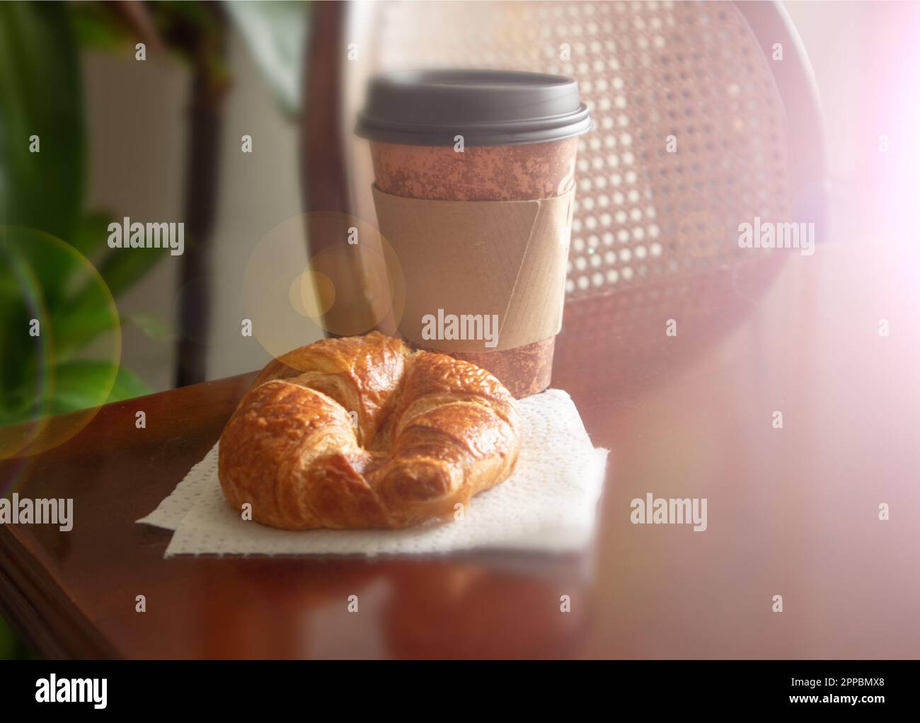 Auf einem Holztisch mit Hintergrundbeleuchtung stehen eine Tasse Kaffee und ein Croissant aus Pappe zur Verfügung. Stockfoto