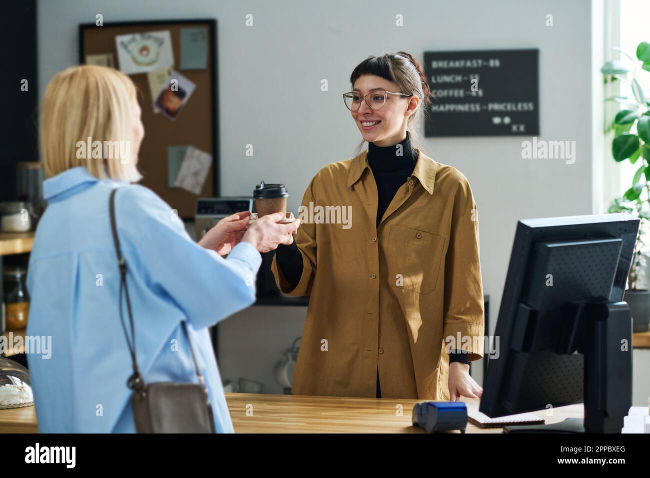 Junge Kellnerin mit Brille, die blonde, reife Kundin mit einem Lächeln ansieht, während sie ihre Tasse Kaffee über den Tresen im Café verteilt Stockfoto