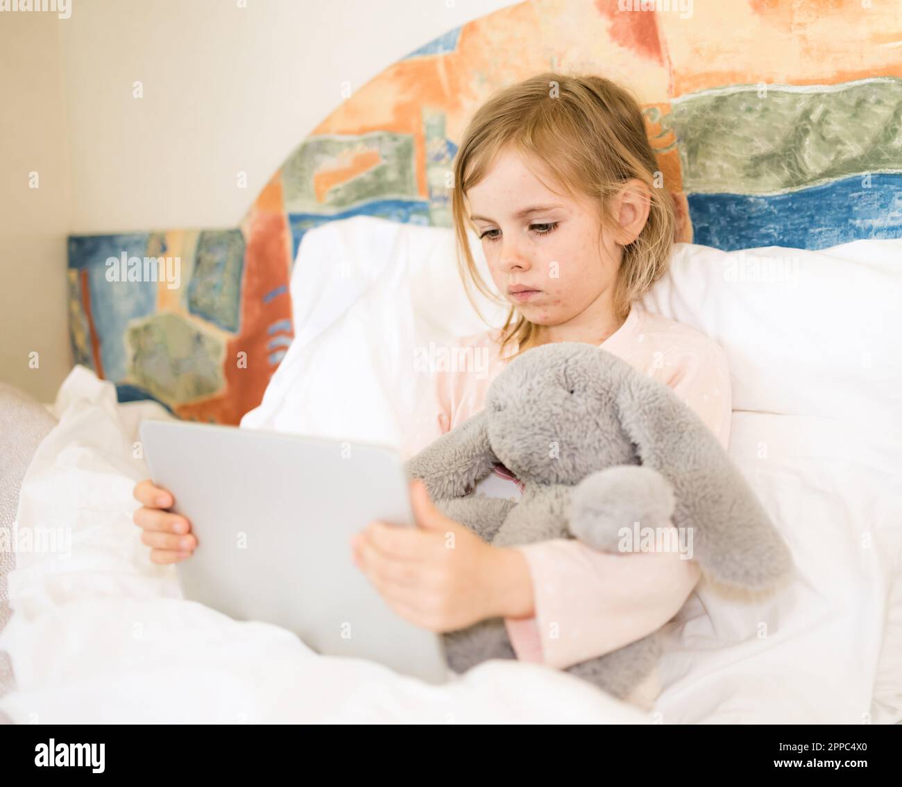 Krankes Kind mit roten Pickel, das im Bett sitzt und flauschiges Spielzeug umarmt. Ein kleines Mädchen, das auf ein Tablet schaut. Windpocken, Varizellen-Virus oder vesikulärer Ausschlag bei Kindern Stockfoto